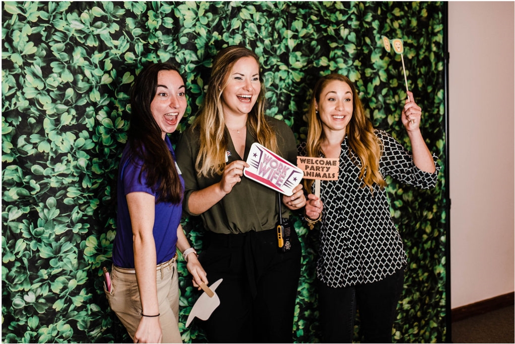 three women pose for a photo booth in front of a faux vine wall
